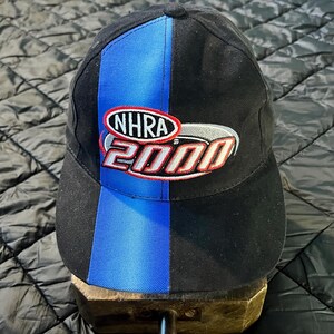 K&ouml;nnte beinhalten: Schwarze Baseballkappe mit blauem vertikalen Streifen und dem gestickten Text "NHRA 2000" in Rot, Wei&szlig; und Silber. Die Kappe hat einen gebogenen Schirm und einen Knopf oben.