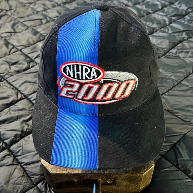 K&ouml;nnte beinhalten: Schwarze Baseballkappe mit blauem vertikalen Streifen und dem gestickten Text "NHRA 2000" in Rot, Wei&szlig; und Silber. Die Kappe hat einen gebogenen Schirm und einen Knopf oben.