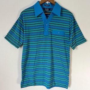 May include: A short-sleeved polo shirt with horizontal stripes in shades of blue, teal, and black. The shirt features a solid turquoise collar, a three-button placket, and a chest pocket. The shirt is hanging on a wooden hanger.