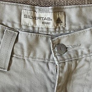 May include: Close-up of khaki-coloured trousers with the brand label "SILVERTAB" visible. The trousers feature a button closure and a zip. The fabric has a textured appearance.