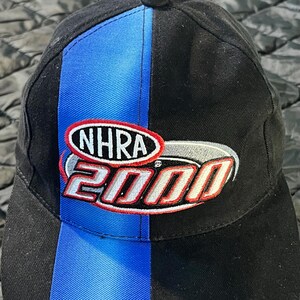 K&ouml;nnte beinhalten: Schwarze Baseballkappe mit blauem vertikalen Streifen auf der Vorderseite. Die Kappe zeigt das NHRA 2000 Logo in roter, wei&szlig;er und silberner Stickerei. Die Kappe ist aus strapazierf&auml;higem Stoff gefertigt.