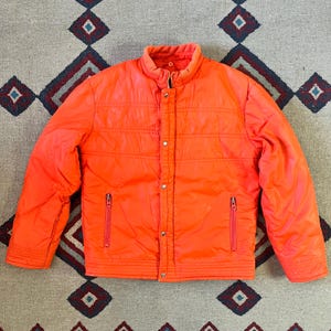May include: Bright orange quilted jacket with a stand-up collar, snap and zipper closures, and two front pockets. The jacket has a ribbed hem and cuffs. The jacket is displayed on a patterned rug.