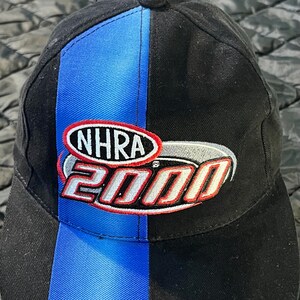 K&ouml;nnte beinhalten: Schwarze Baseballkappe mit blauem vertikalen Streifen und dem NHRA 2000 Logo. Das Logo ist rot, wei&szlig; und silber. Die Kappe ist aus strapazierf&auml;higem Stoff und hat einen gebogenen Schirm.