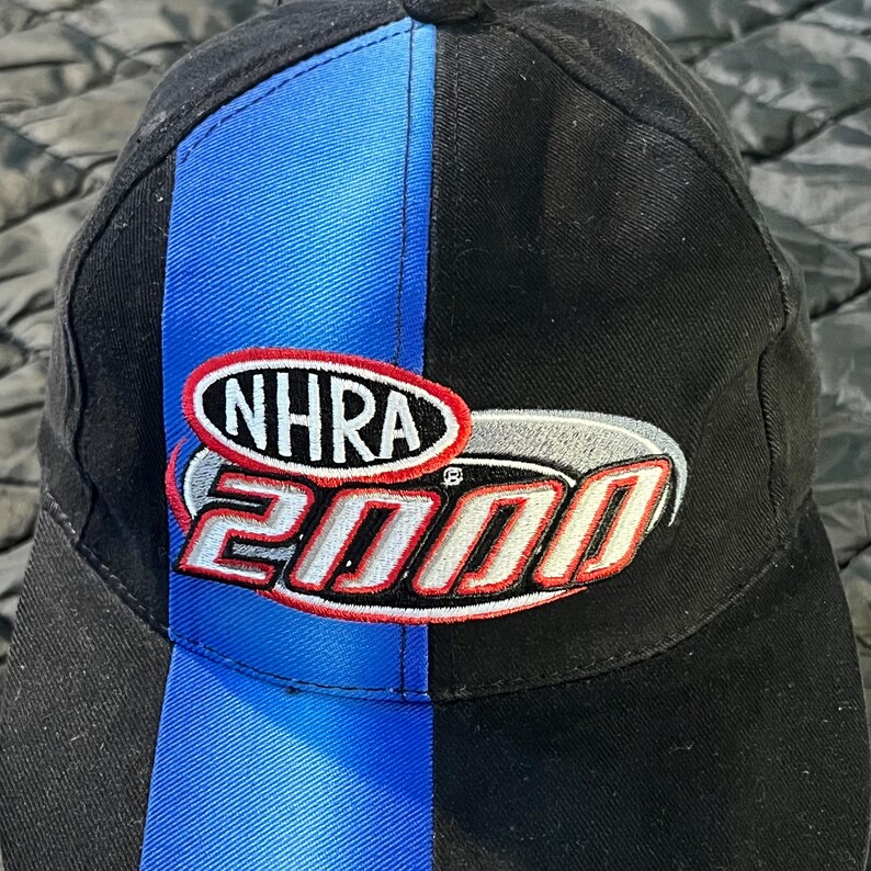 K&ouml;nnte beinhalten: Schwarze Baseballkappe mit blauem vertikalen Streifen und dem NHRA 2000 Logo. Das Logo ist rot, wei&szlig; und silber. Die Kappe ist aus strapazierf&auml;higem Stoff und hat einen gebogenen Schirm.