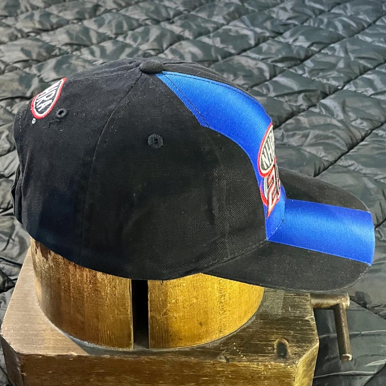 K&ouml;nnte beinhalten: Schwarze Baseballkappe mit blauem Streifen und Logo. Die Kappe hat einen gebogenen Schirm und einen Knopf oben. Das Logo ist wei&szlig; und rot. Die Kappe liegt auf einer Holzform.