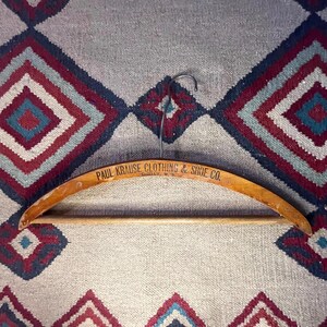 May include: A vintage wooden clothing hanger with the text "PAUL KRAUSE CLOTHING & SHOE CO." in black lettering. The hanger has a curved design and a metal hook. The background is a patterned textile with red, blue, and white diamond shapes.