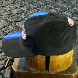 K&ouml;nnte beinhalten: Schwarze Baseballkappe mit blauem Schirm und blauem Einsatz oben. Die Kappe hat ein Logo an der Seite und ein kleines Emblem auf der R&uuml;ckseite. Die Kappe steht auf einem Holzst&auml;nder.
