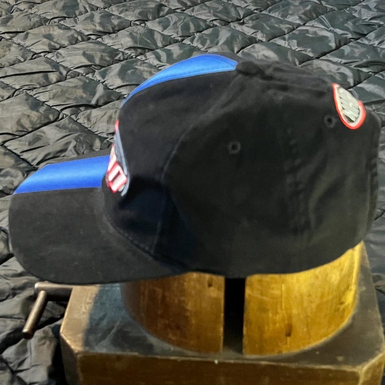 K&ouml;nnte beinhalten: Schwarze Baseballkappe mit blauem Schirm und blauem Einsatz oben. Die Kappe hat ein Logo an der Seite und ein kleines Emblem auf der R&uuml;ckseite. Die Kappe steht auf einem Holzst&auml;nder.