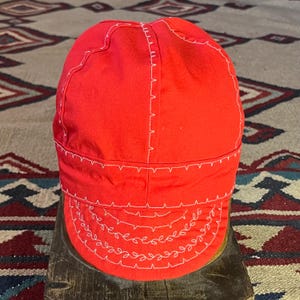 May include: Bright red welder's cap with white stitching. The cap features a curved brim and is constructed with multiple panels. The stitching is visible along the seams and around the brim, creating a decorative element. The cap is designed to protect the wearer from sparks and heat.