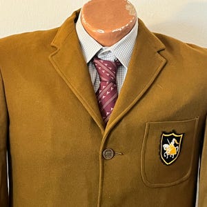 May include: A brown wool blazer with a single button closure and a patch pocket on the right side. The pocket features a gold embroidered emblem of a knight on horseback.
