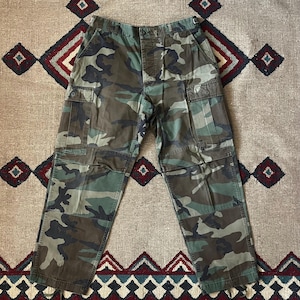 May include: Camouflage cargo pants in shades of green, brown, and black. The pants feature multiple pockets on the legs and a button and zipper closure. The fabric has a military-inspired pattern.