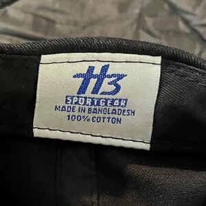 K&ouml;nnte beinhalten: Nahaufnahme einer schwarzen Baseballkappe mit einem wei&szlig;en Etikett. Das Etikett zeigt das blaue Logo "HB" und den Text "SPORTGEAR MADE IN BANGLADESH 100% COTTON". Die Kappe ist aus Baumwolle.