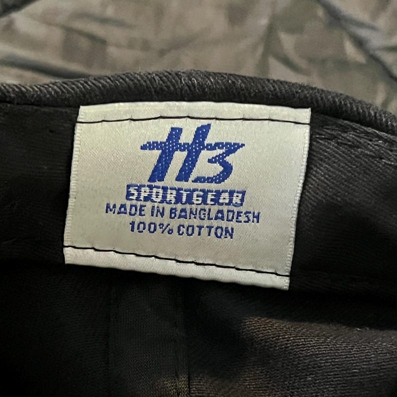 K&ouml;nnte beinhalten: Nahaufnahme einer schwarzen Baseballkappe mit einem wei&szlig;en Etikett. Das Etikett zeigt das blaue Logo "HB" und den Text "SPORTGEAR MADE IN BANGLADESH 100% COTTON". Die Kappe ist aus Baumwolle.