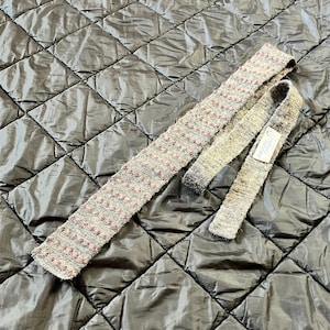 Puede incluir: Una corbata gris y roja estampada con una correa estrecha a juego. La corbata tiene un aspecto texturizado con pequeños puntos rojos. Una etiqueta blanca con texto azul está adherida a la correa estrecha.