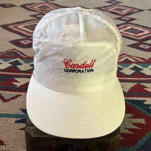 Peut inclure: Casquette de baseball blanche avec une visière incurvée. Le chapeau affiche le texte rouge "Cardell" au-dessus du texte noir "CORPORATION". La casquette est faite d'un matériau léger et texturé et possède un bouton sur le dessus.