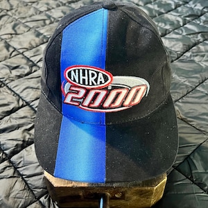 K&ouml;nnte beinhalten: Schwarze Baseballkappe mit einem vertikalen blauen Streifen und dem Text "NHRA 2000" in Rot, Wei&szlig; und Silber. Die Kappe hat einen gebogenen Schirm und ist wahrscheinlich aus strapazierf&auml;higem Stoff gefertigt.