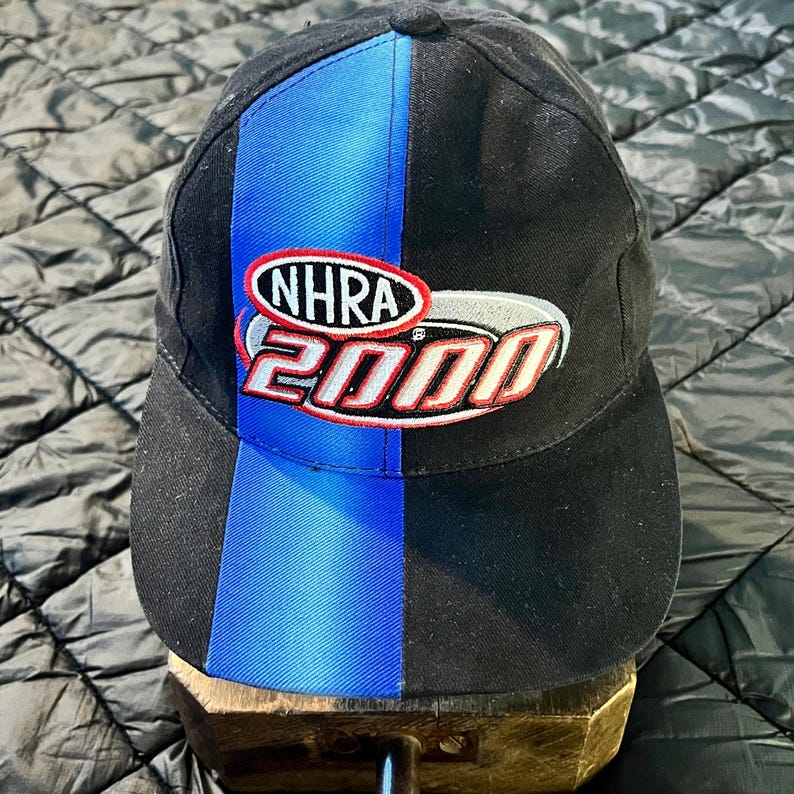 K&ouml;nnte beinhalten: Schwarze Baseballkappe mit einem vertikalen blauen Streifen und dem Text "NHRA 2000" in Rot, Wei&szlig; und Silber. Die Kappe hat einen gebogenen Schirm und ist wahrscheinlich aus strapazierf&auml;higem Stoff gefertigt.