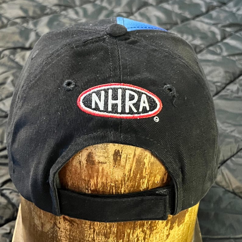 K&ouml;nnte beinhalten: Schwarze Baseballkappe mit blauem Schirm und einem rot-wei&szlig;-schwarzen NHRA-Logo. Die Kappe hat einen gebogenen Schirm und einen verstellbaren Riemen. Hergestellt aus strapazierf&auml;higem Material, geeignet f&uuml;r Outdoor-Aktivit&auml;ten.