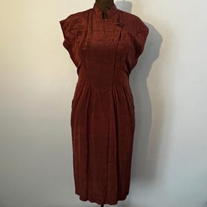 May include: A maroon sleeveless dress with a mandarin collar and a single button closure at the neck. The dress has a fitted bodice and a slightly flared skirt.
