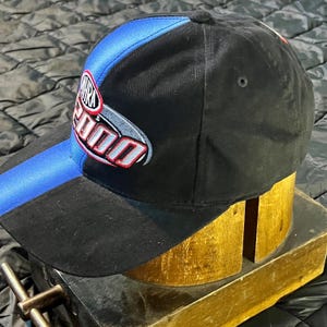 K&ouml;nnte beinhalten: Schwarze Baseballkappe mit blauem Streifen auf dem Schirm und einem Logo mit der Aufschrift "NHRA 2000" in Rot, Wei&szlig; und Silber. Die Kappe wird auf einer h&ouml;lzernen Hutform pr&auml;sentiert.
