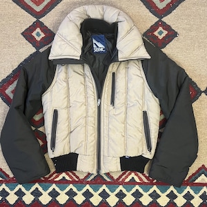 May include: A beige and black quilted winter jacket with a high collar and a full-length zipper. The jacket has black sleeves and a black ribbed hem. The brand name "SERAC" is visible on a tag inside the collar.