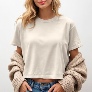May include: Cream-colored, cropped t-shirt with short sleeves. A beige cardigan is draped over the shoulders. The model is wearing blue jeans.