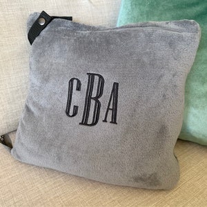 K&ouml;nnte beinhalten: Ein graues Pl&uuml;sch-Wurfkissen mit einem schwarzen Riemen und einem schwarzen gestickten Monogramm "CBA".