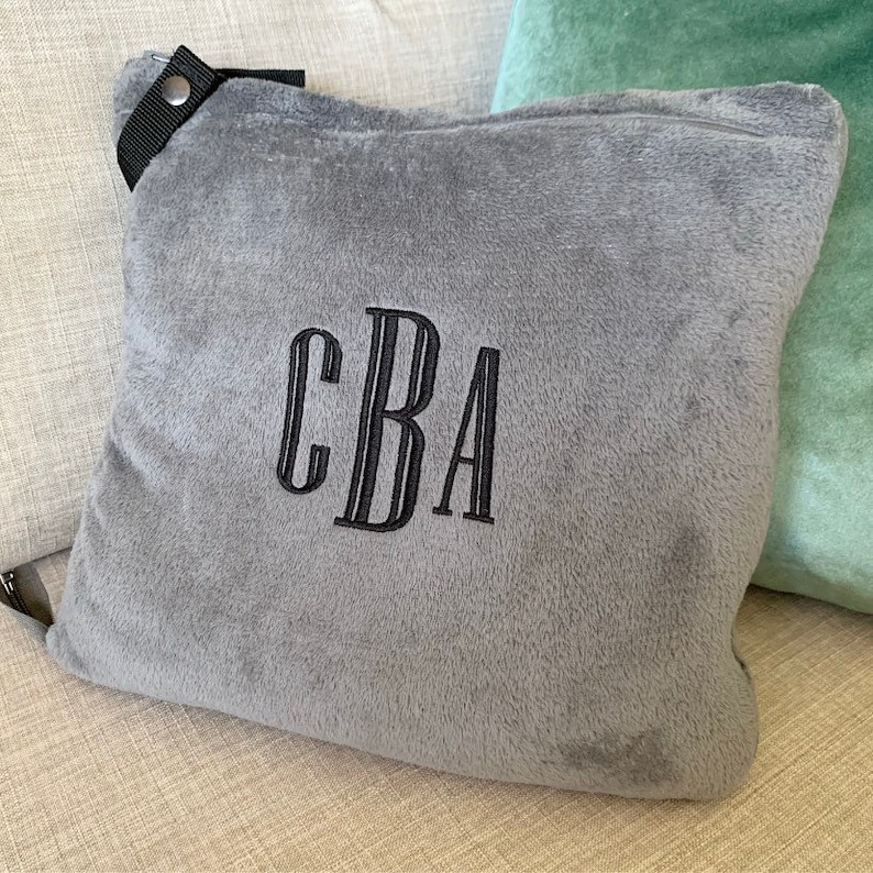 K&ouml;nnte beinhalten: Ein graues Pl&uuml;sch-Wurfkissen mit einem schwarzen Riemen und einem schwarzen gestickten Monogramm "CBA".