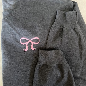 Könnte beinhalten: Ein dunkelgraues Langarmshirt mit einer aufgestickten rosa Schleife auf der Vorderseite. Das Shirt ist in der Hälfte gefaltet und die Ärmel sind hochgekrempelt.