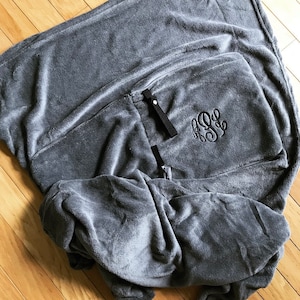 K&ouml;nnte beinhalten: Eine graue Fleece-Decke mit einem schwarzen gestickten Monogramm. Die Decke hat zwei schwarze B&auml;nder.