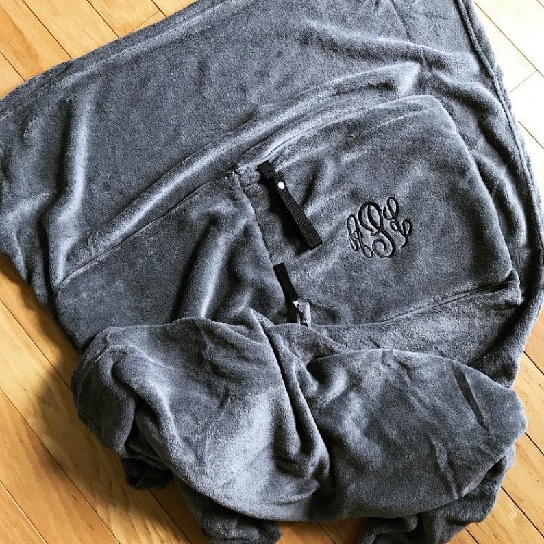 K&ouml;nnte beinhalten: Eine graue Fleece-Decke mit einem schwarzen gestickten Monogramm. Die Decke hat zwei schwarze B&auml;nder.