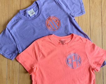 Puede incluir: Dos camisetas con monogramas bordados. La camiseta de arriba es de color morado claro con el monograma "ATB" en naranja. La camiseta de abajo es de color naranja brillante con el monograma "EPK" en azul.