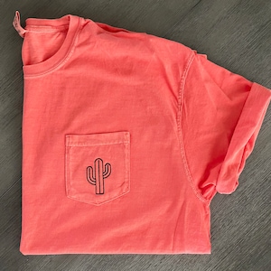 Puede incluir: Una camiseta rosa coral con un gráfico de cactus negro en un bolsillo.