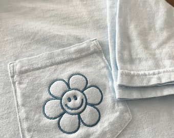 Może przedstawiać: Jasno błękitny t-shirt z naszywaną kieszenią, na której znajduje się haftowana niebieska stokrotka z uśmiechniętą buzią.