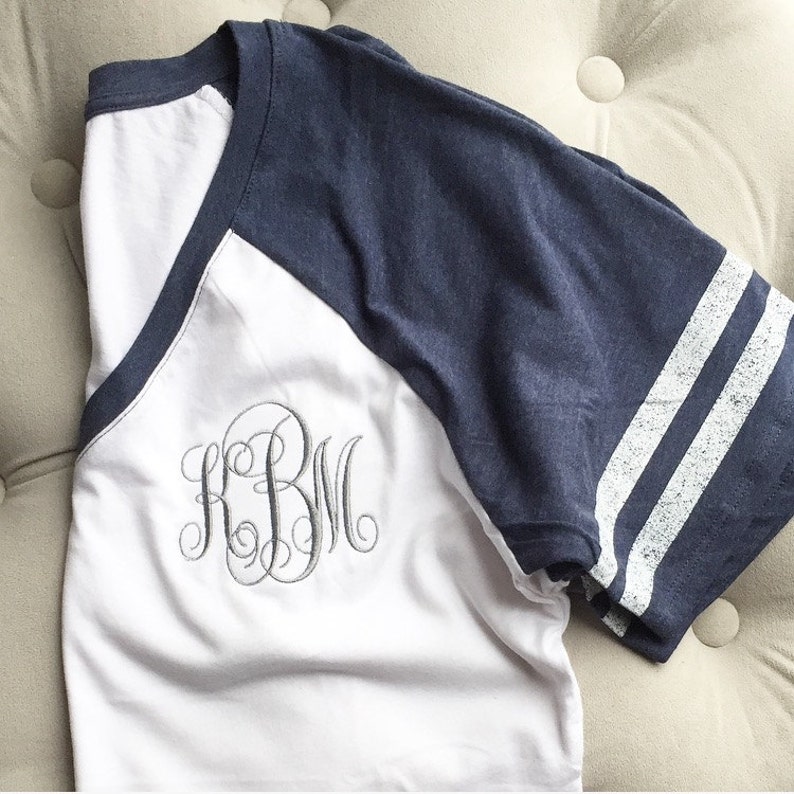 Puede incluir: Una camiseta blanca con una manga de estilo b&eacute;isbol azul marino y blanca. La camiseta tiene cuello en V y un monograma de las letras "KBM" bordado en la parte delantera.