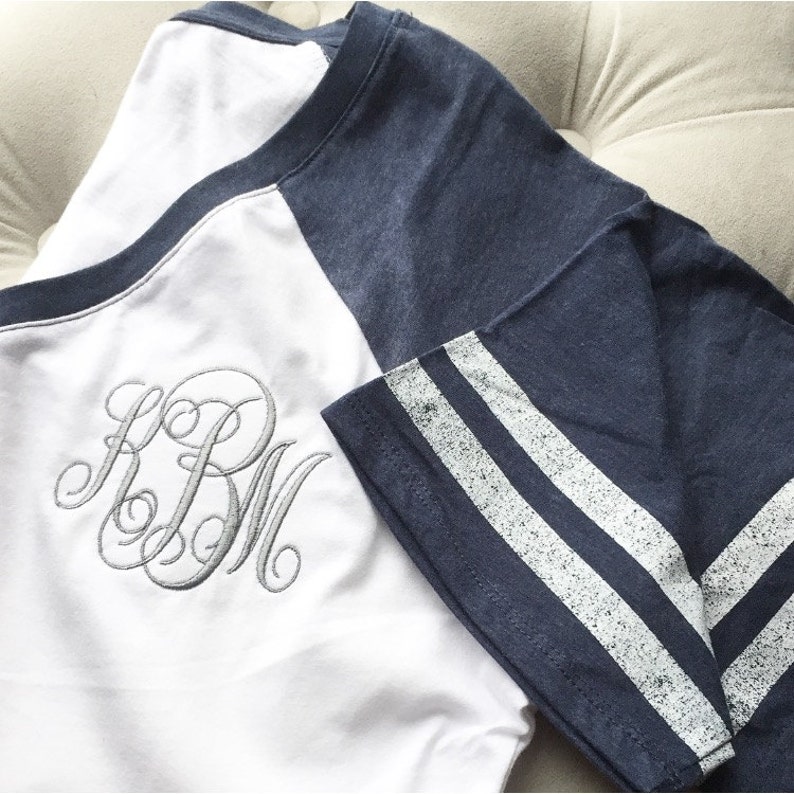 Puede incluir: Una camiseta de b&eacute;isbol blanca y azul marino con rayas blancas en las mangas. La camiseta tiene un monograma "KBM" bordado en la parte delantera en hilo gris.