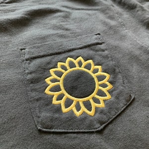 May include: A black t-shirt with a pocket featuring an embroidered yellow sunflower design.