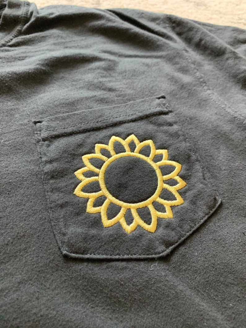 May include: A black t-shirt with a pocket featuring an embroidered yellow sunflower design.