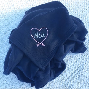 May include: A navy blue fleece blanket with a pink heart and arrow design embroidered on it. The heart has the name "Mia" inside.