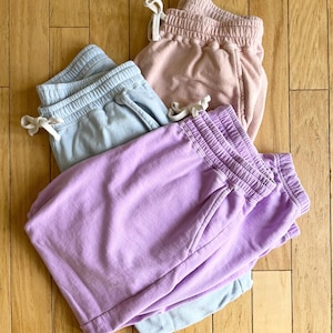Może przedstawiać: Cztery pary casualowych szortów dresowych w pastelowych kolorach: jasnoniebieskim, różowym i fioletowym. Każda para ma talię ze sznurkiem i kieszenie. Szorty są wykonane z miękkiego materiału i ułożone na drewnianej powierzchni.