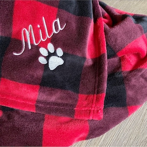 May include: Red and black plaid fleece blanket with the name "Mila" embroidered in white with a white paw print below it.