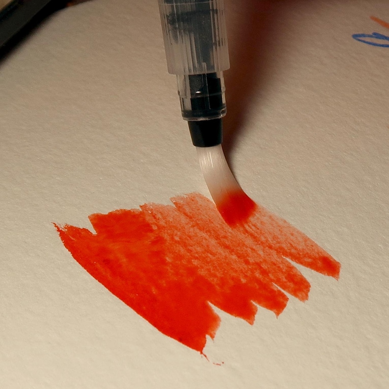 Puede incluir: Primer plano de un bol&iacute;grafo pincel con una punta blanca, creando un trazo de acuarela roja sobre papel blanco.