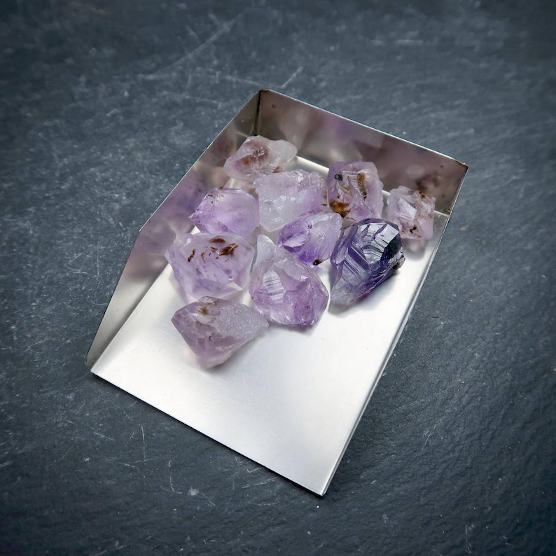 May include: A collection of raw amethyst crystals in a silver scoop. The crystals vary in size and shape, displaying shades of purple and lavender with some brown inclusions. The scoop is positioned on a dark gray surface.