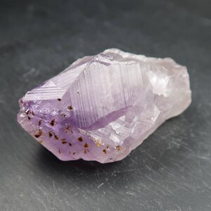 May include: A raw amethyst crystal with a light purple hue and a crystalline structure. The gemstone has a rough, natural texture with small, dark inclusions and a few golden specks. The crystal is approximately 3 inches long.
