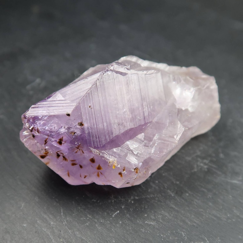 May include: A raw amethyst crystal with a light purple hue and a crystalline structure. The gemstone has a rough, natural texture with small, dark inclusions and a few golden specks. The crystal is approximately 3 inches long.