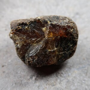 May include: A rough, irregularly shaped amber stone with a dark brown exterior and translucent, golden-brown interior. The surface has a textured appearance, with visible cracks and imperfections. The stone is likely a natural gemstone or mineral specimen.