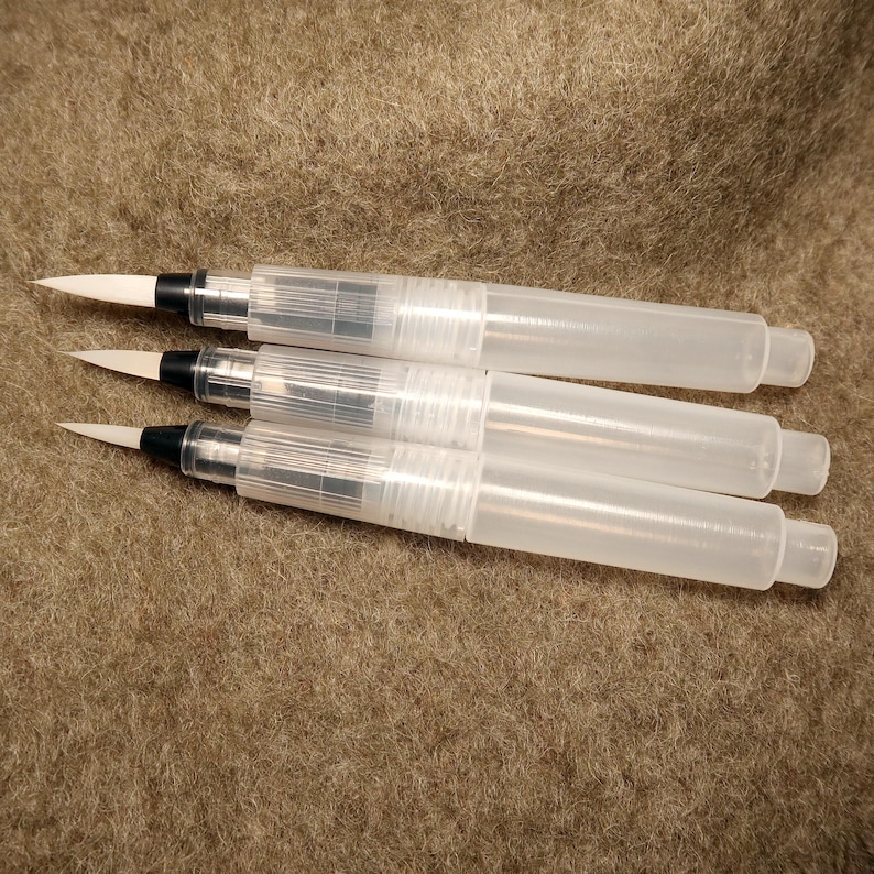 Puede incluir: Tres bol&iacute;grafos de pincel de acuarela de pl&aacute;stico transparente con cerdas blancas. Los bol&iacute;grafos est&aacute;n sobre una superficie marr&oacute;n y peluda.