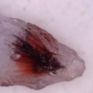 May include: Close-up of a clear quartz crystal with a dark red and black inclusion. The crystal has a rough, natural texture and is set against a white background. The red inclusion appears feathery.