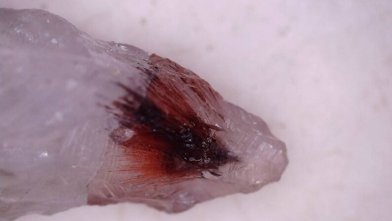 May include: Close-up of a clear quartz crystal with a dark red and black inclusion. The crystal has a rough, natural texture and is set against a white background. The red inclusion appears feathery.