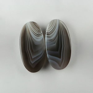 May include: Two oval-shaped agate cabochons with a banded pattern in shades of grey and white.